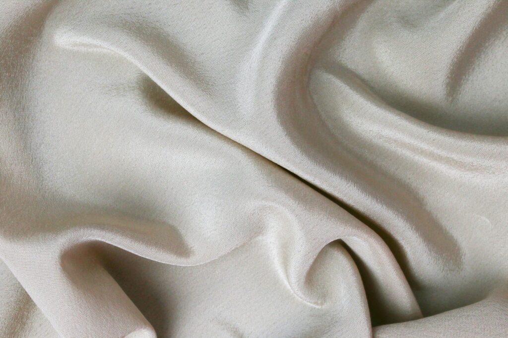 Close-up of ivory silk fabric lying in soft rolling folds, with a low sheen running along the ridges and deeper shadow in the troughs