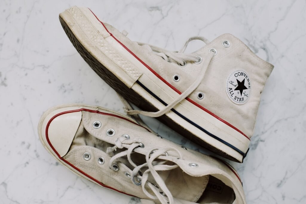 A pair of grubby cream canvas high-top sneakers on white marble seen from above, one tipped to show a dirt-marked rubber outsole and striped midsole, the other with loose laces and a scuffed toe cap
