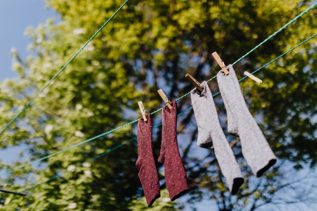 Four socks pegged to a green washing line against sunlit tree branches, two burgundy patterned dress socks and two thick grey marled socks hanging to air dry