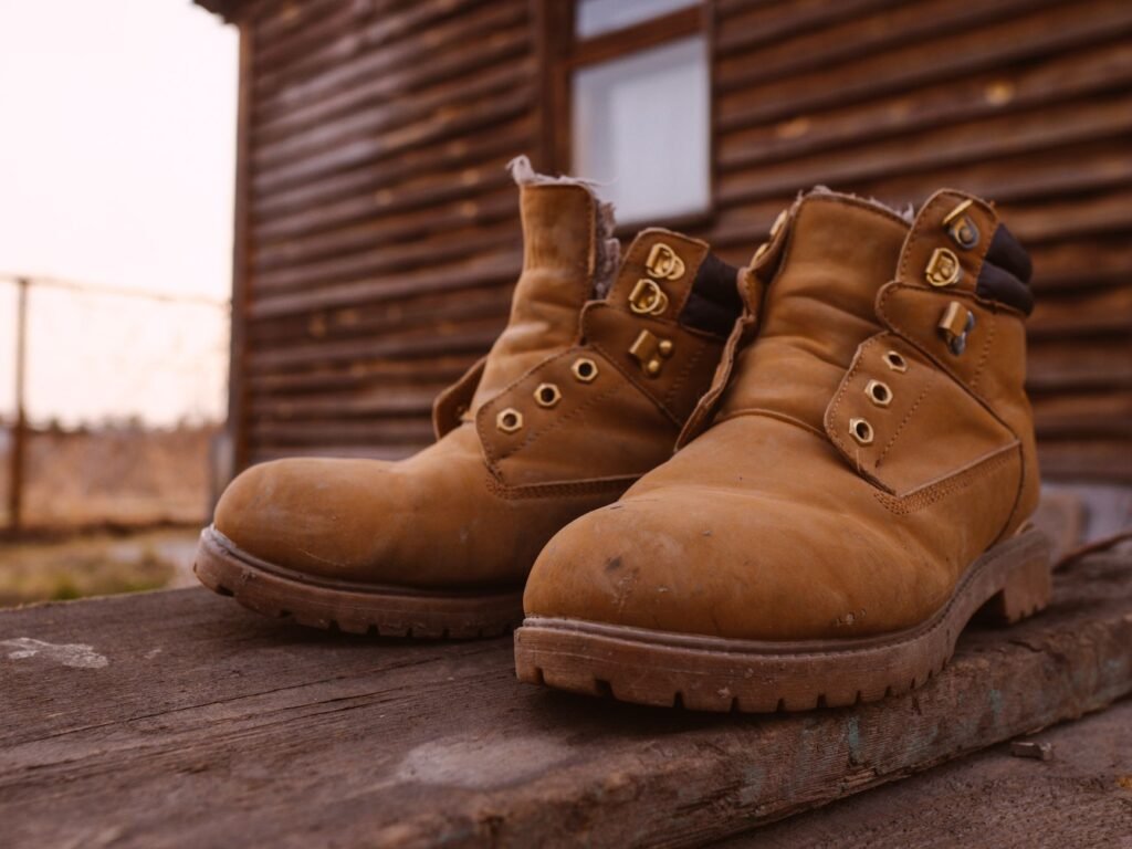 Pair of worn tan suede work boots with the laces removed, standing dusty on a weathered plank outside a log cabin