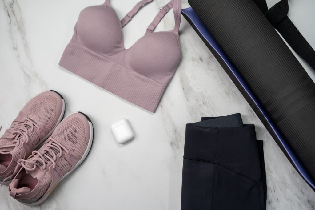 Flat lay on marble of a pink sports bra, pink running shoes, folded navy leggings, a rolled dark yoga mat and a pair of wireless earbuds