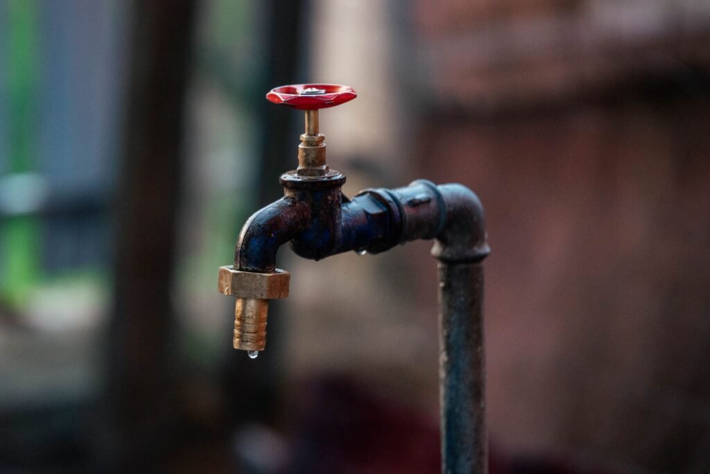 A weathered dark iron outdoor tap with a brass hose adaptor and a single drop forming at the spout