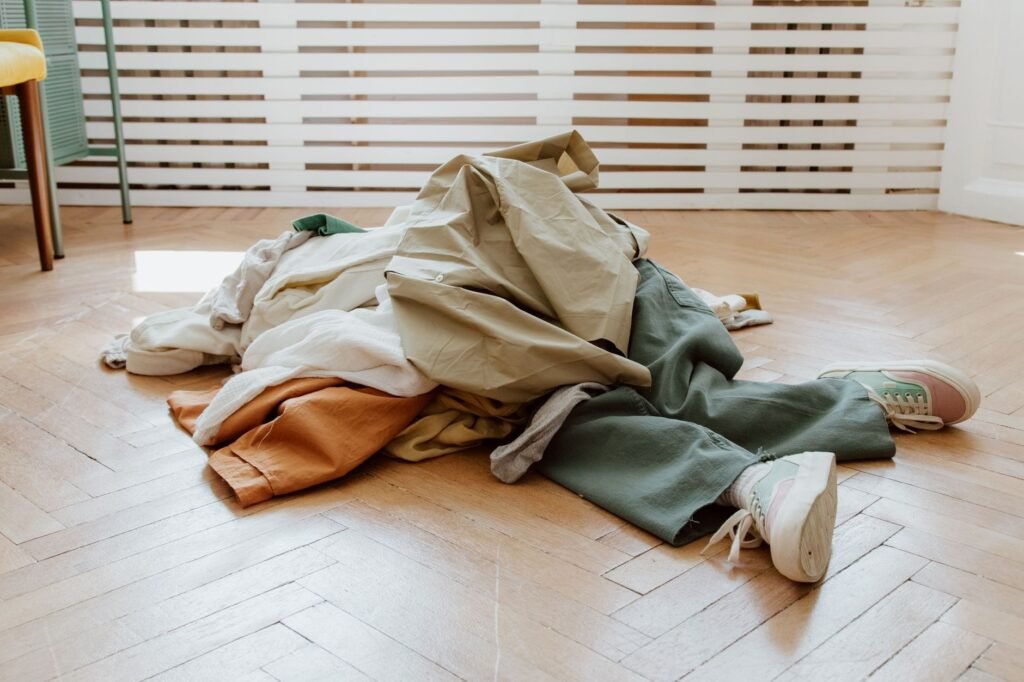 A heap of unfolded laundry on a herringbone parquet floor, pale shirts over orange and olive trousers with grey socks and a pair of trainers beside it