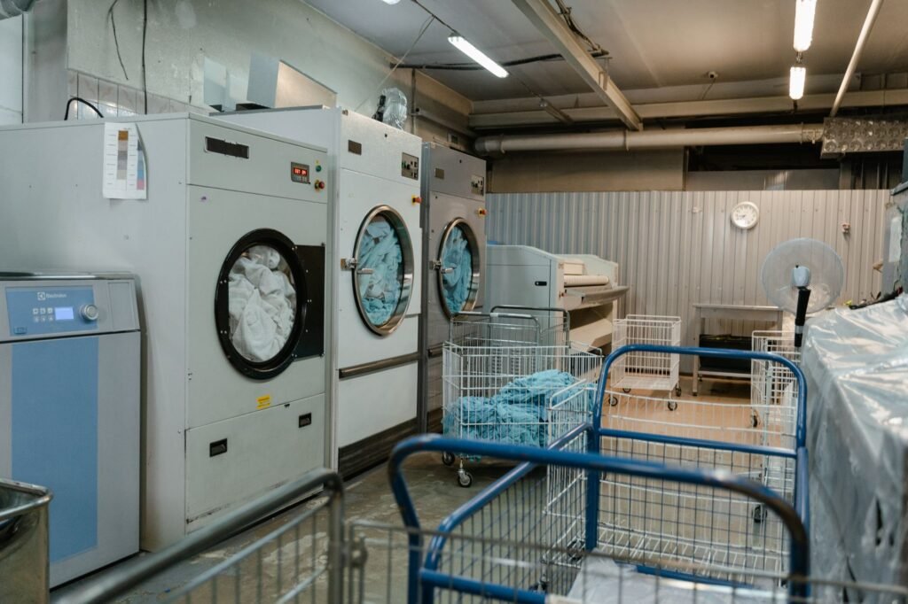 Wire laundry carts on swivel casters standing on a wet concrete floor in front of three large commercial front-load machines, the working arrangement a laundry cart on wheels is copied from