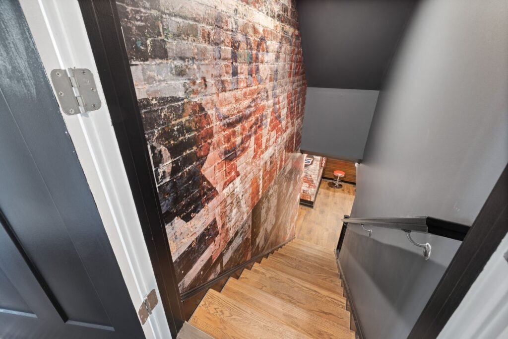 Looking down an open dark interior door at a timber staircase descending to a lower floor, with a painted brick feature wall alongside it