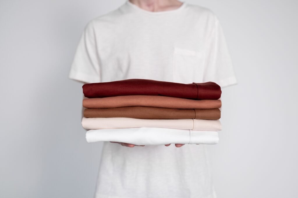 A person in a white t-shirt holding a stack of five folded garments in dark red, rust, tan, cream and white against a plain pale grey wall