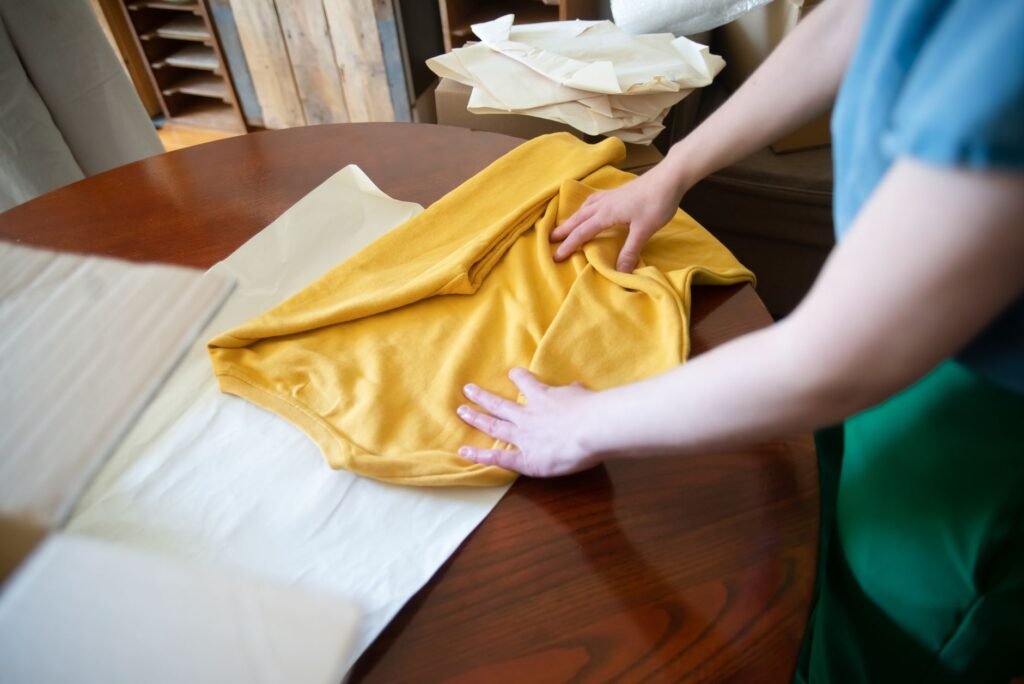 Two hands smoothing a folded mustard-yellow sweatshirt flat on a round dark wood table beside a sheet of pale tissue paper