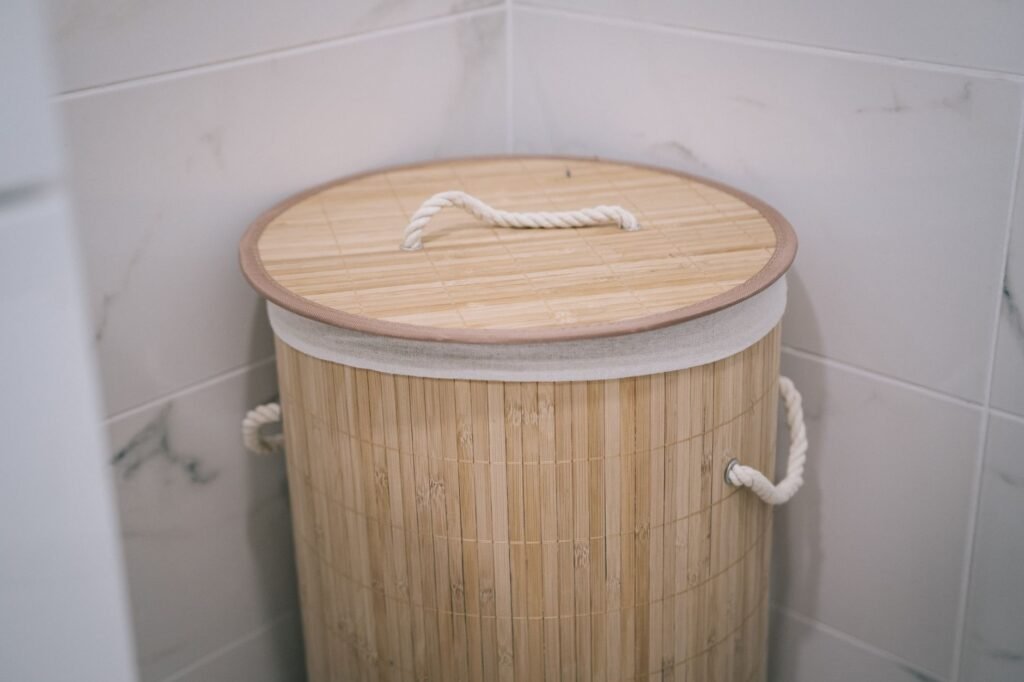 A round bamboo slat laundry hamper with a fitted bamboo lid and white rope handles, standing in the corner of a white marble-tiled bathroom