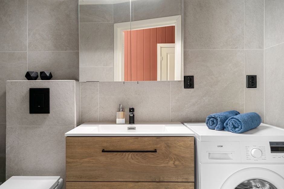 A white front-load washing machine with rolled blue towels on top, standing beside a wood vanity drawer unit in a grey tiled bathroom
