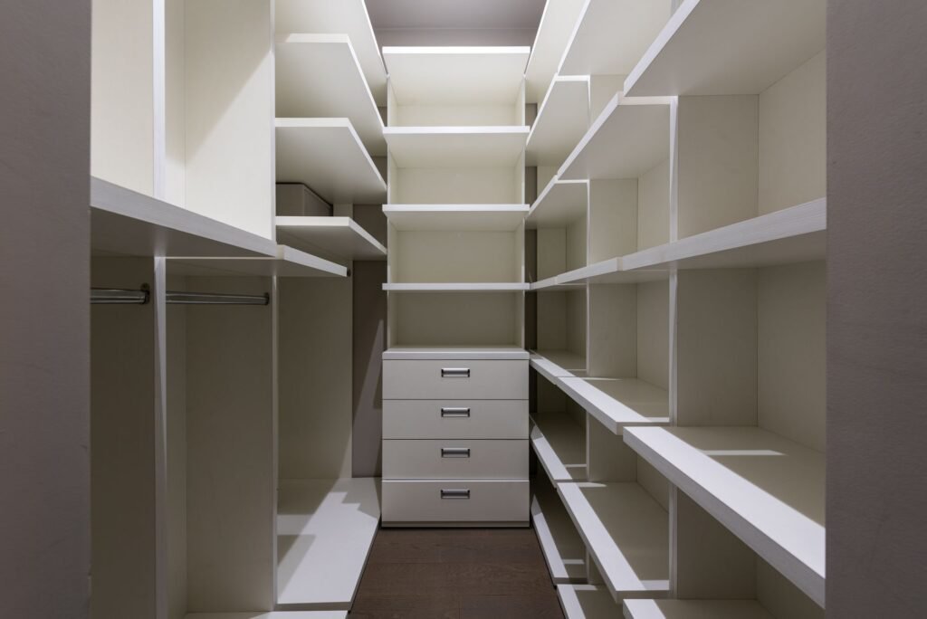 An empty fitted storage room lined with white shelves on both sides, a steel hanging rail on the left and a four-drawer unit at the far end