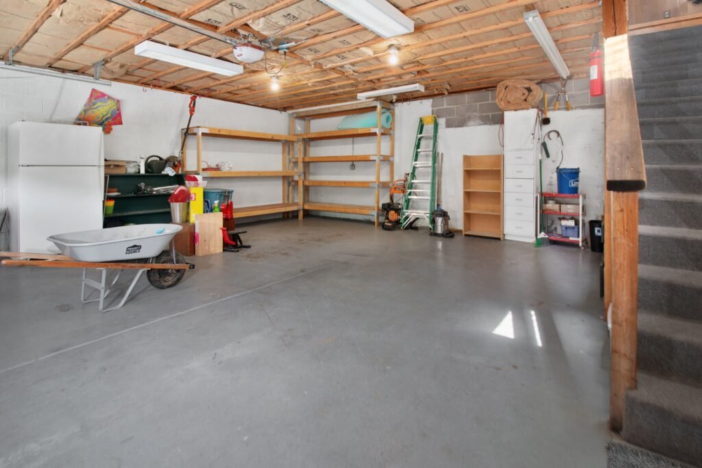 An unfinished garage with exposed ceiling joists, painted concrete block walls, timber shelving, a white chest freezer, a wheelbarrow on a bare concrete floor and carpeted stairs on the right