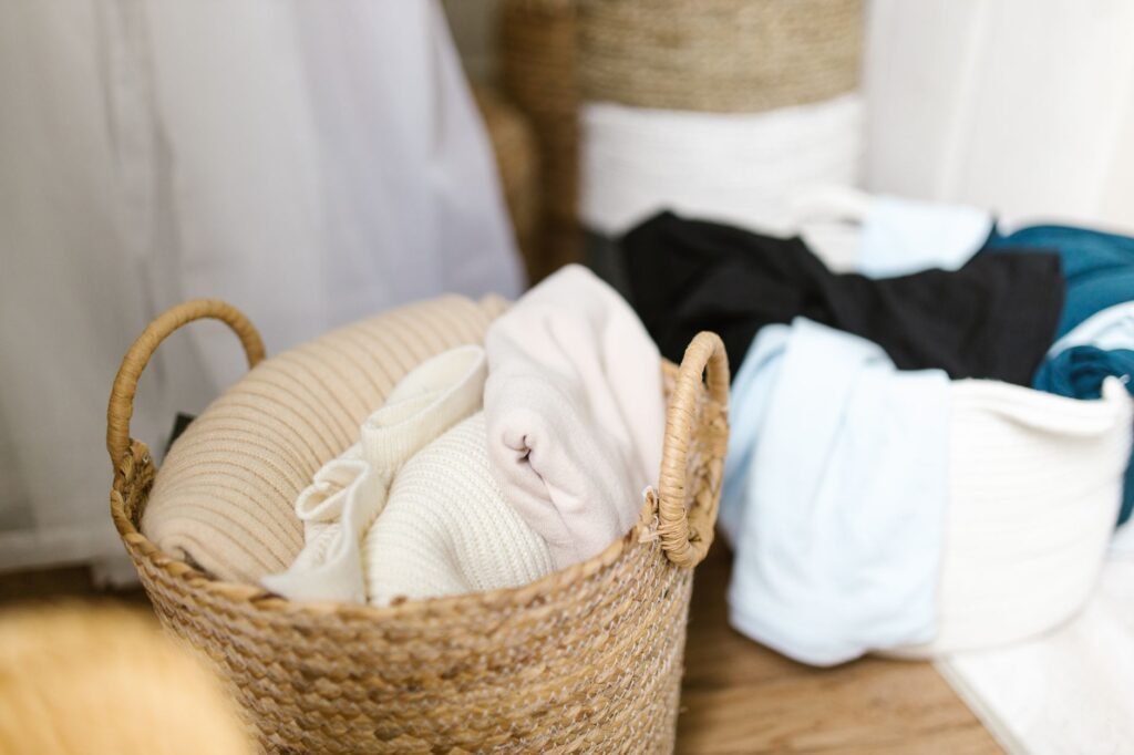 A woven seagrass basket holding folded cream and oatmeal knitwear beside a white rope basket of blue and black clothing on a wooden floor