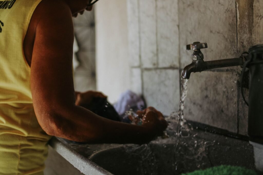 A person in a yellow vest hand-washing a dark garment in a deep concrete utility basin with a wall-mounted tap running, the soaking and hand-washing job a laundry tub sink exists for