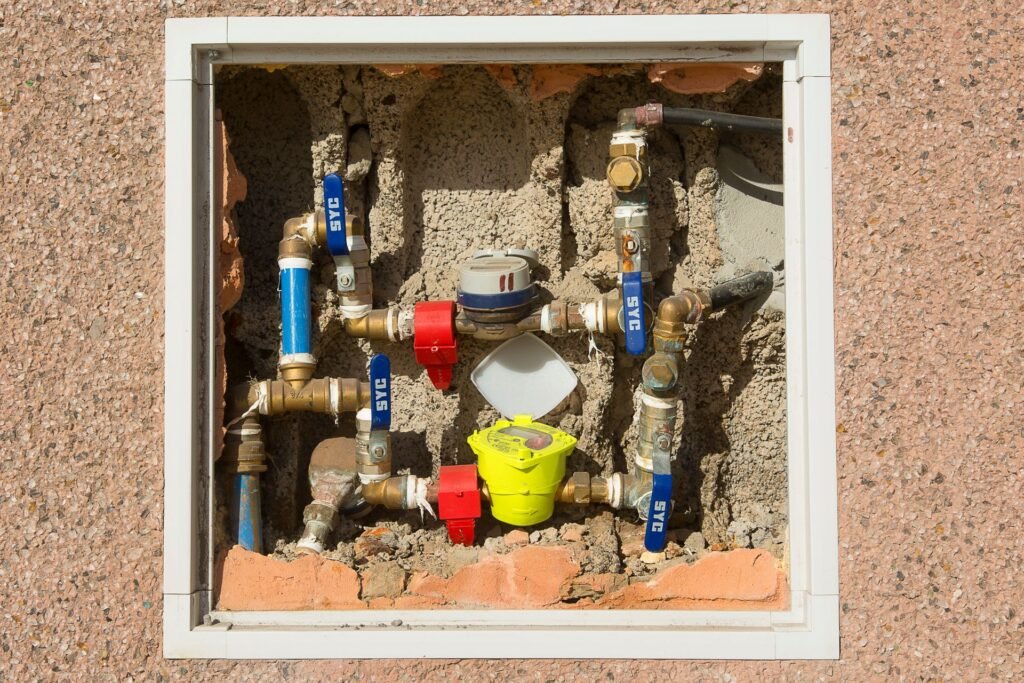 A recessed wall cabinet holding two domestic water meters, brass unions and blue-handled ball valves, the point in the plumbing where an automatic shutoff valve is fitted