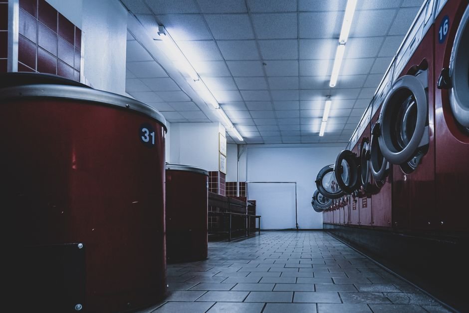 A dim laundromat under fluorescent strip lights with a row of dark red front-load machines and their still, empty chrome-rimmed drums along the right wall