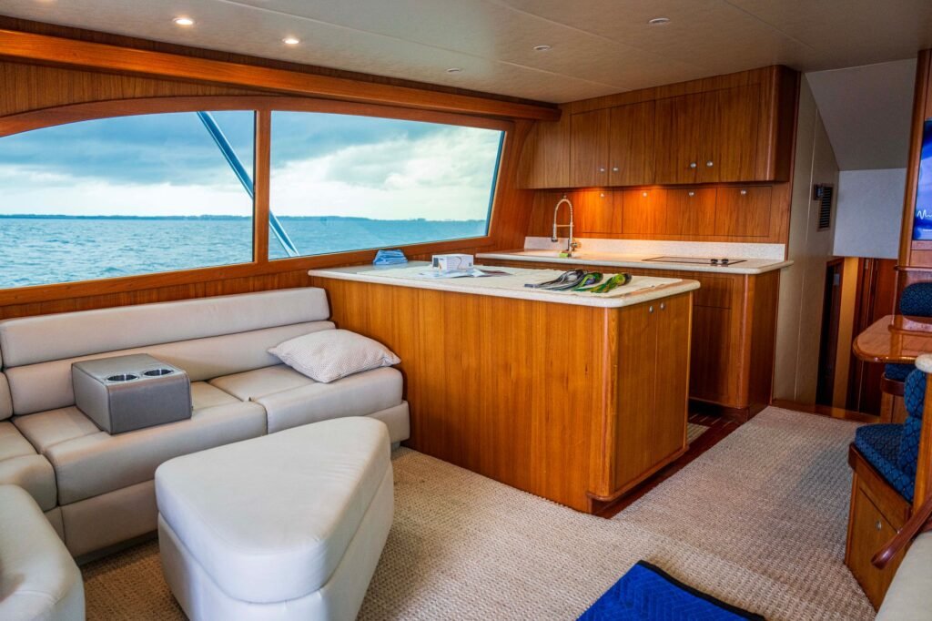 Teak-lined saloon of a motor yacht with a galley counter, sink and induction hob beside a leather settee and a wide window onto open water, the space a marine washing machine competes for