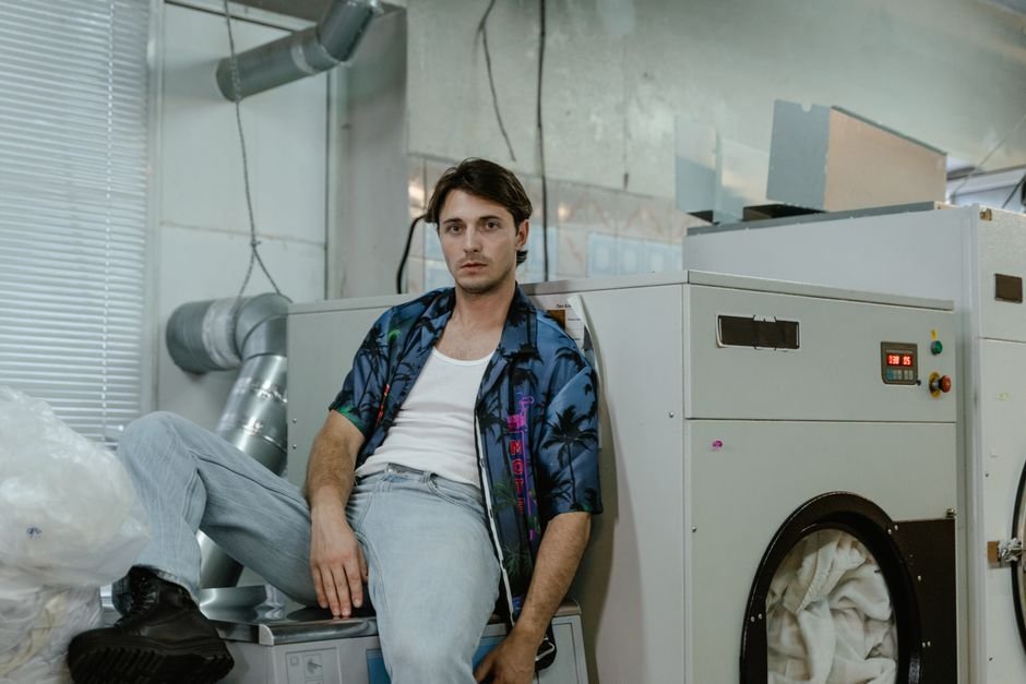 A man seated in a commercial laundry beside a tumble dryer holding white bedding, its control panel showing a red digital readout, with wide silver flexible exhaust ducting running along the wall behind him