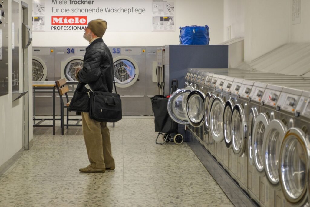 An older man with a shoulder bag standing in a self-service laundrette, reading a sign above a bank of numbered dryers with a long row of stainless coin-operated front-loaders beside him