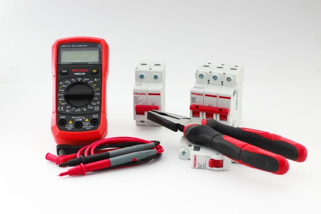 Red digital multimeter marked 600V CAT III beside coiled test leads, miniature circuit breakers, pliers and two screwdrivers on a white background