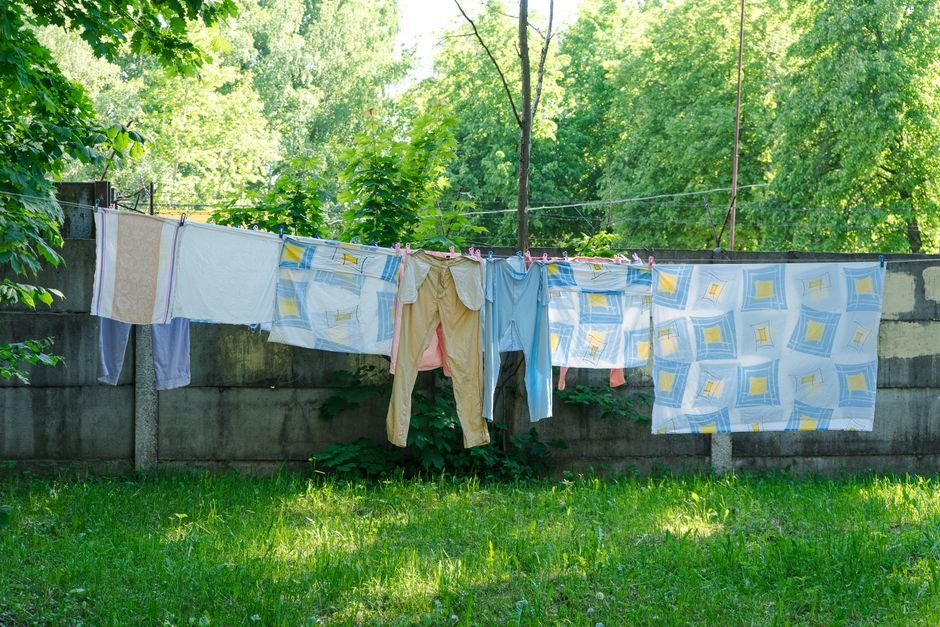 Two parallel washing lines in a sunlit green garden carrying a patterned duvet cover, khaki trousers and pale towels above a low concrete fence