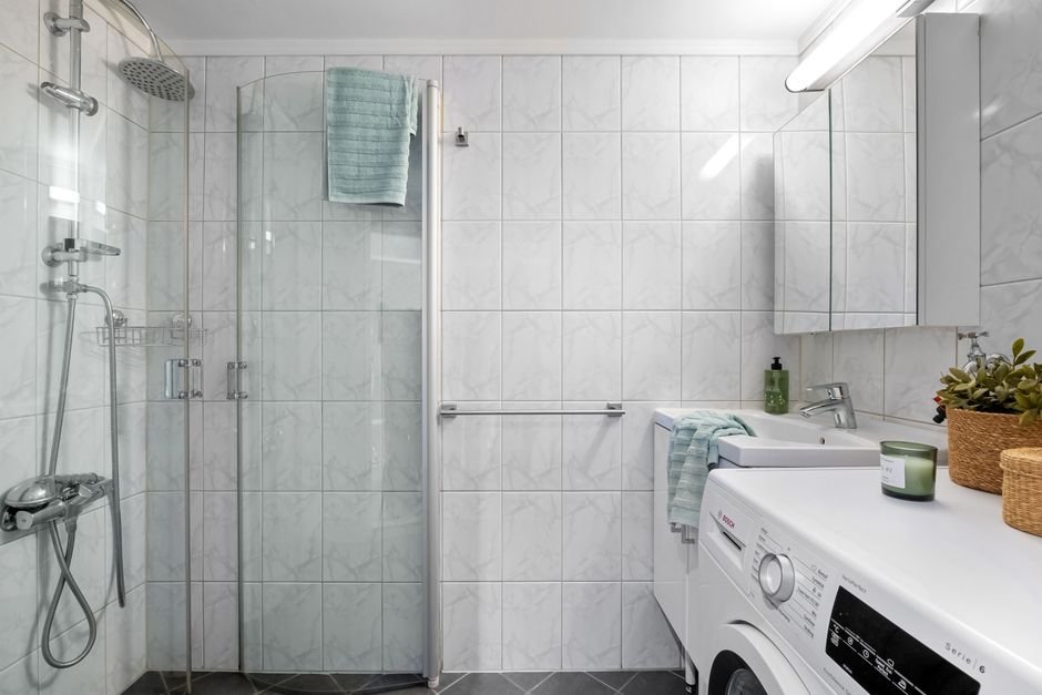 White front-load washing machine built in under a worktop beside a basin, with a mirrored wall cabinet and a folded towel on a rail above the machine