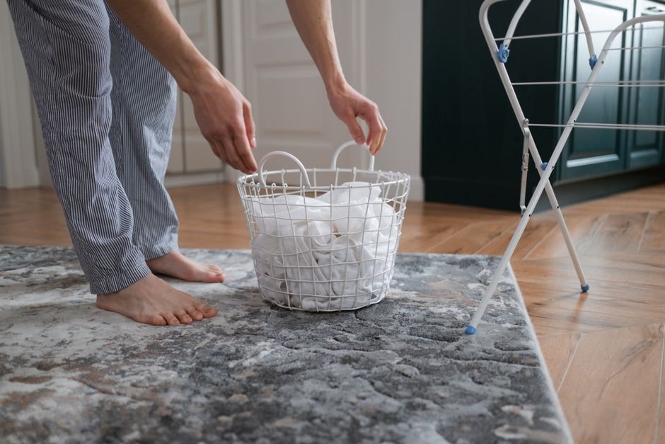 A barefoot person in striped pyjama trousers lifting a white wire basket of wet white washing off a rug, with a folding drying rack standing on the wooden floor behind