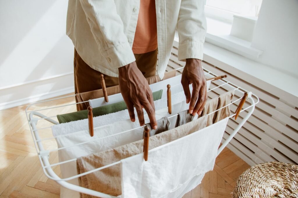 A person arranging damp laundry over the bars of a white folding drying rack in a sunlit room, wooden pegs along the rails, a radiator and parquet floor behind