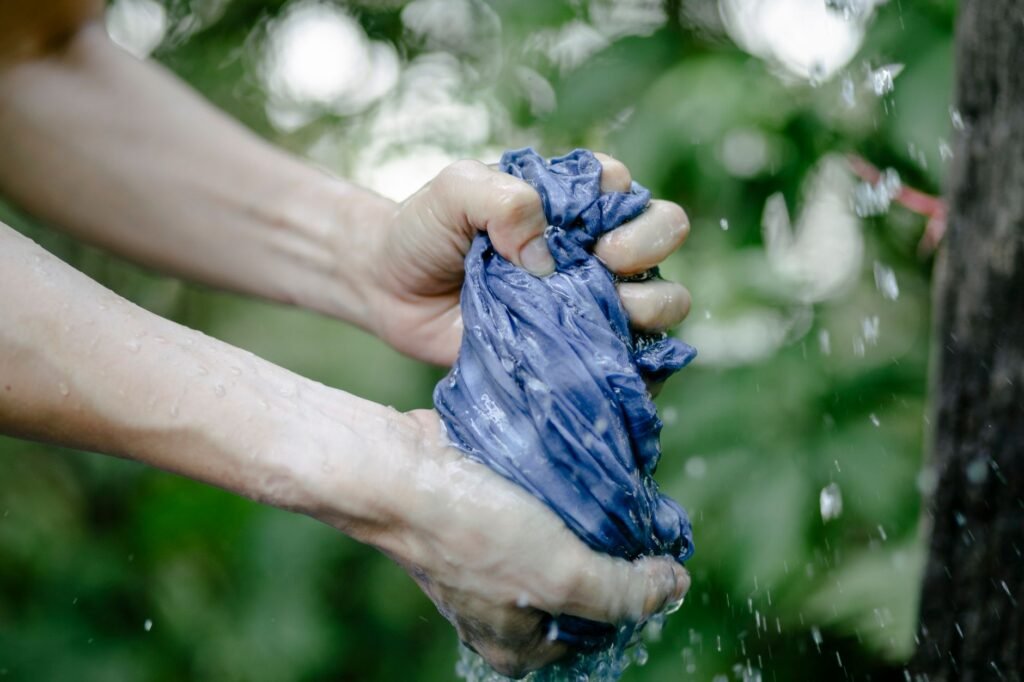 Two bare hands twisting a soaking blue cloth outdoors so water sprays out of it, against a blurred green background