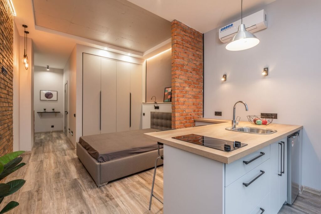 A small studio apartment with a bed beside a short kitchen counter holding a sink and hob, and a narrow corridor leading off to one side