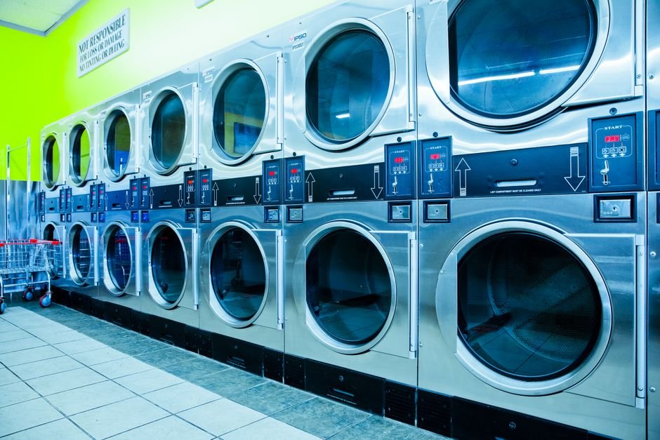 A long bank of stacked stainless steel coin-operated laundromat dryers against a lime green wall, with a wire laundry cart at the end of the row