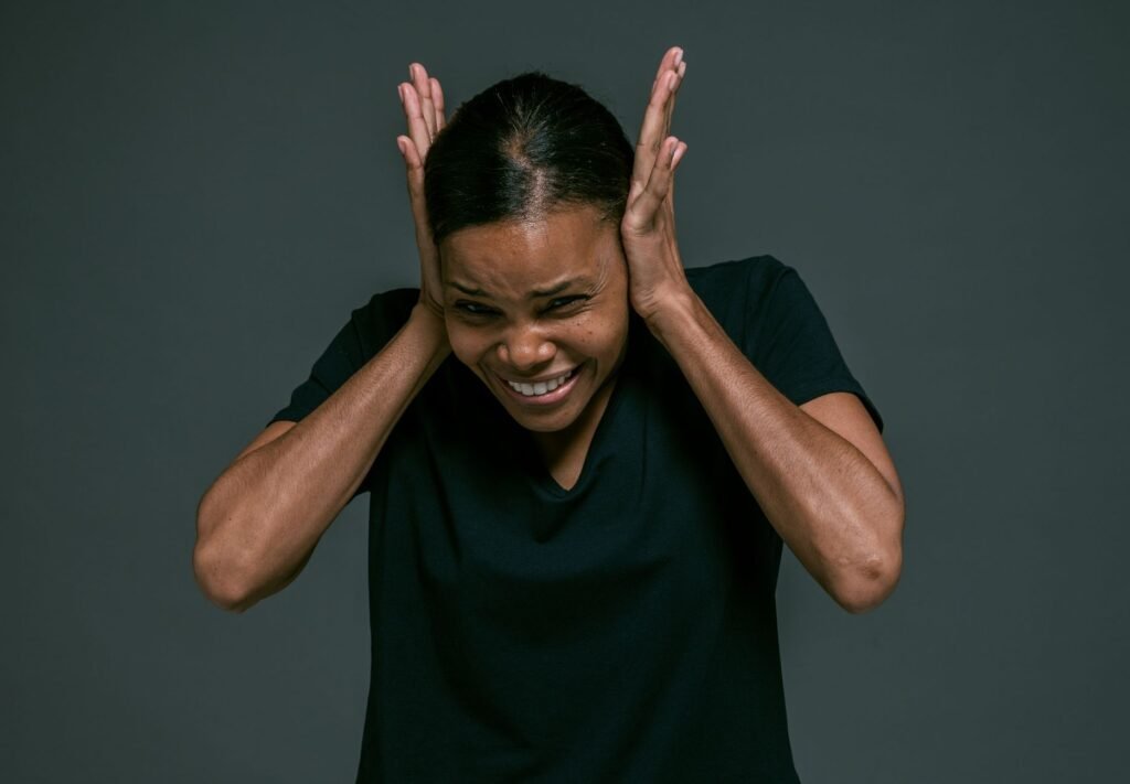 A woman in a black v-neck t-shirt pressing both hands flat over her ears and wincing, photographed against a dark grey studio background
