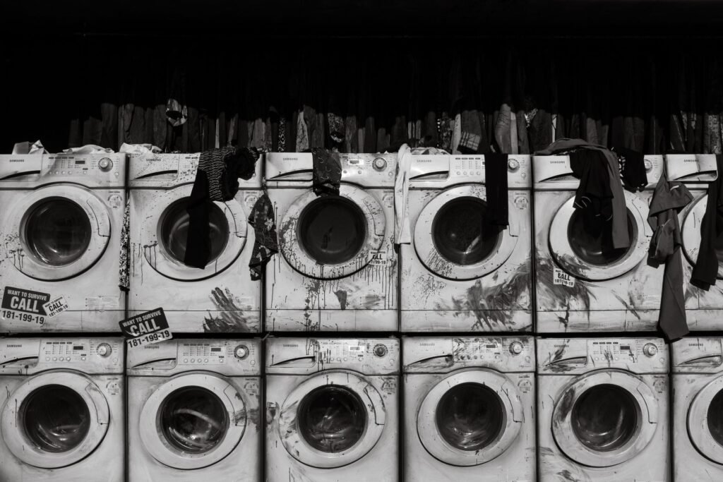 A black-and-white wall of stacked, heavily stained second-hand front-load washing machines with clothes hanging out of some of the doors