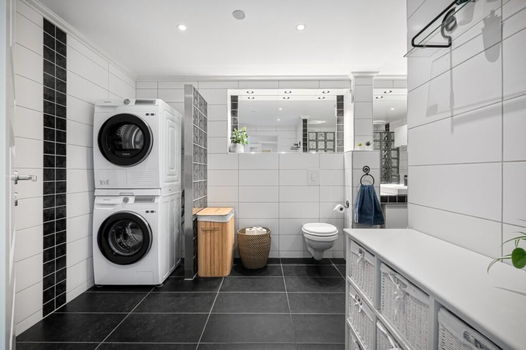 A Samsung heat pump dryer stacked on a Samsung front-load washer in a white-tiled bathroom, both control panels within reach
