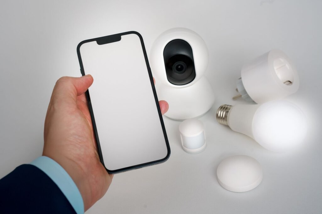 A hand holding a smartphone with a blank screen beside a white smart camera, a smart plug, a lit smart bulb and a motion sensor on a plain white surface