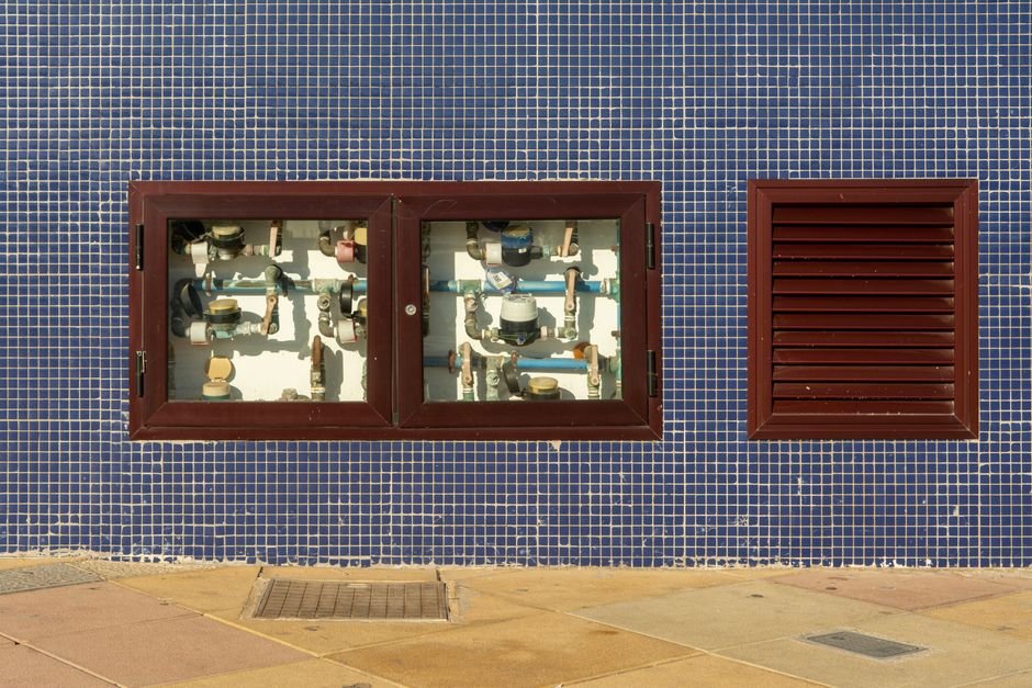 An open utility cabinet in a blue tiled wall exposing a bank of domestic water meters and brass pipework, the metering point a smart water monitor reads from