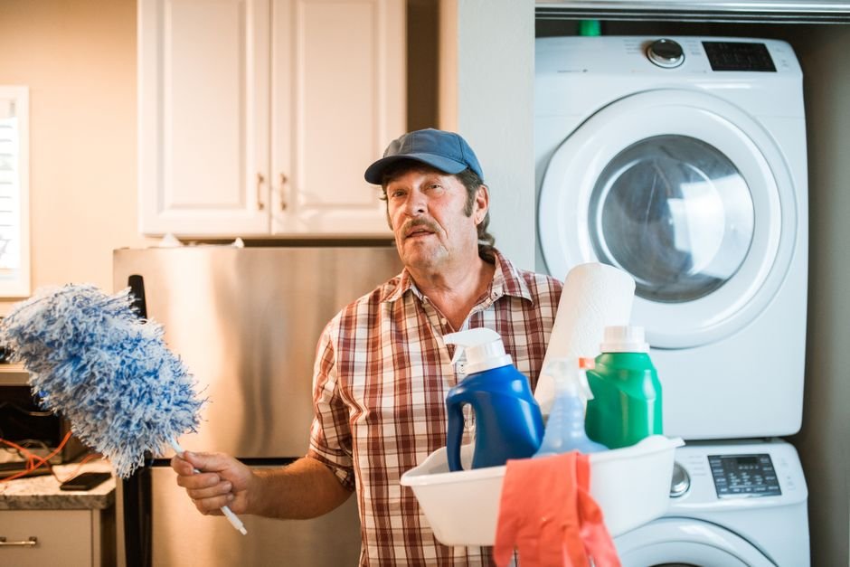 A white dryer stacked directly above a white washing machine in a home laundry alcove, with a man holding a caddy of cleaning supplies beside it