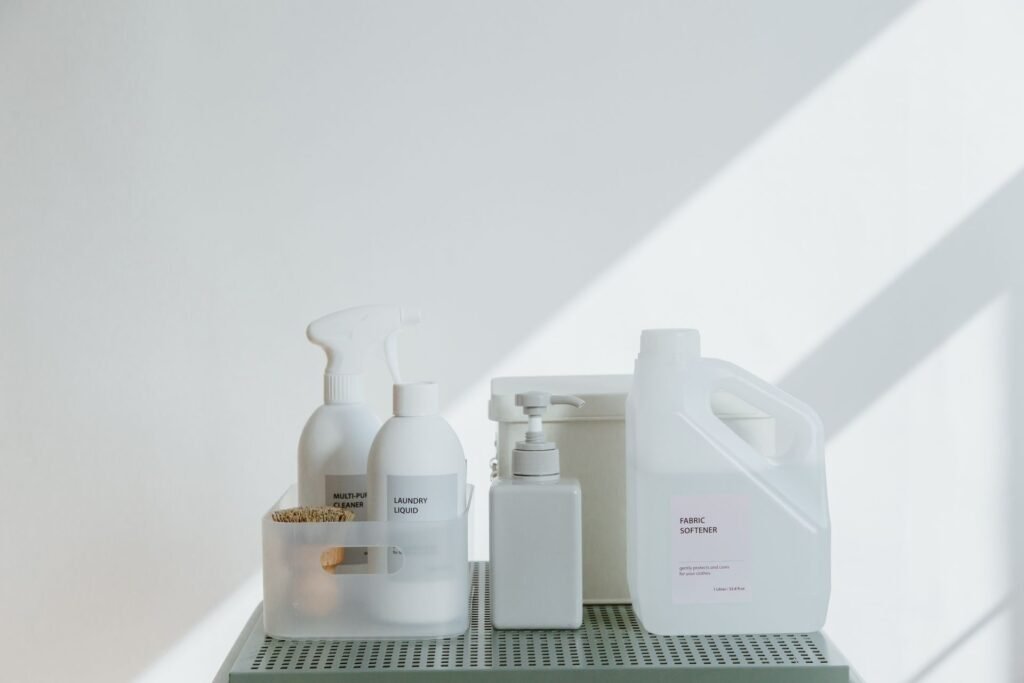 Unbranded white bottles labelled multi-purpose cleaner, laundry liquid and fabric softener standing on a pale green rack in hard sunlight