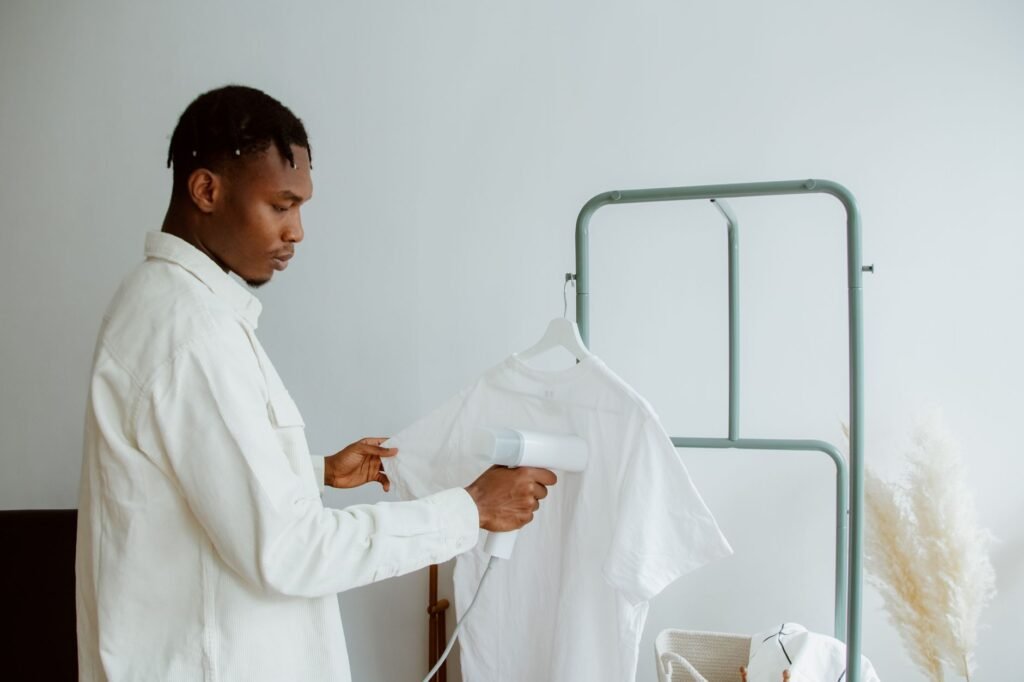A man holds the hem of a white T-shirt taut on a green clothes rail while running a small steamer head up the fabric