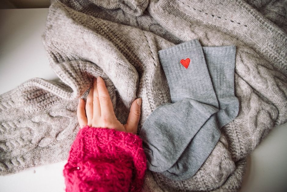 Grey socks and a hand in a pink knitted cuff resting on folded cable-knit jumpers, photographed from above on a white surface