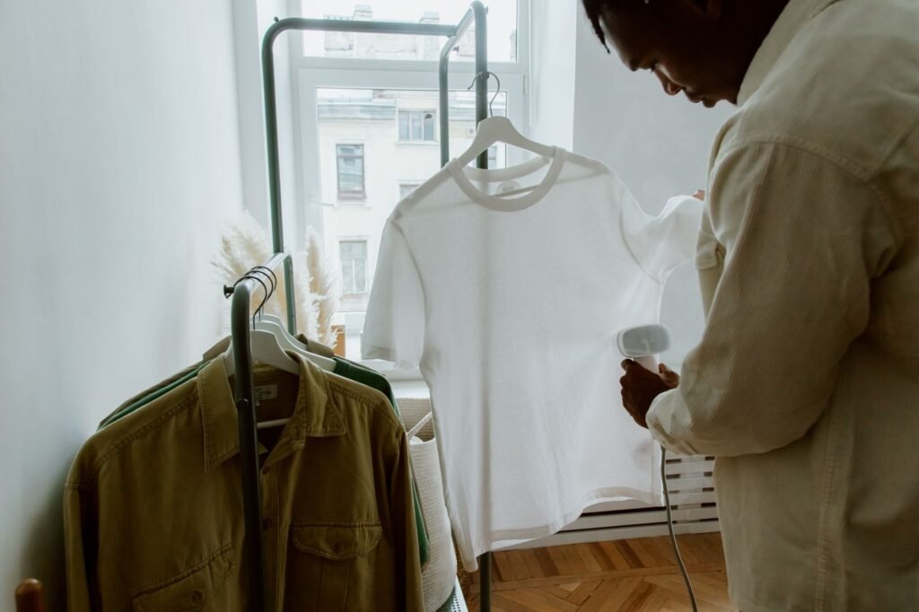 A man in a cream shirt holding a handheld garment steamer against a white t-shirt hanging on a clothes rail by a window, with olive shirts on a lower rail