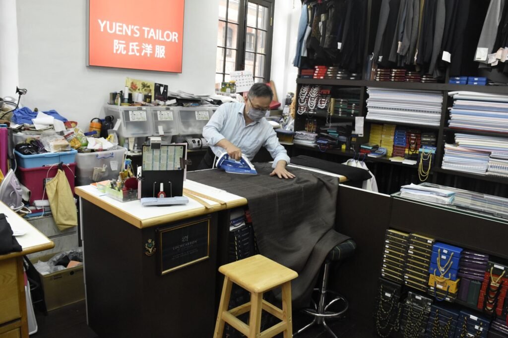 A tailor presses a long flat length of dark cloth spread across a table in a garment shop lined with pattern books and hanging suits