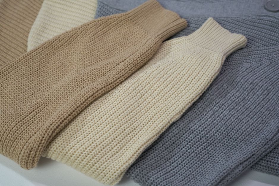 Three ribbed knit sweaters in tan, cream and grey laid out flat side by side with their sleeves extended on a white surface