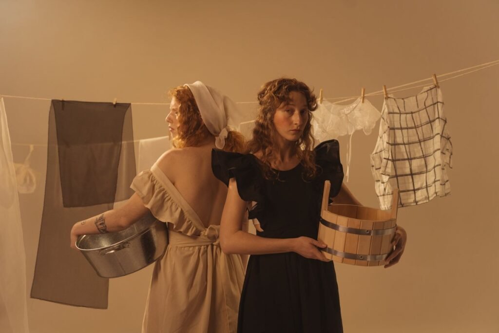 Two people standing back to back, one holding a metal basin and the other a wooden tub, in front of a washing line hung with clothes