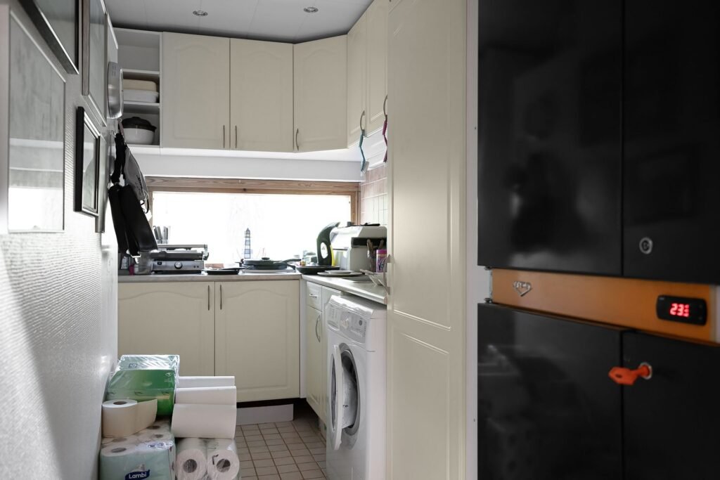 A narrow galley kitchen with a front-load washing machine built in under the worktop, wall cabinets and a window above the counter