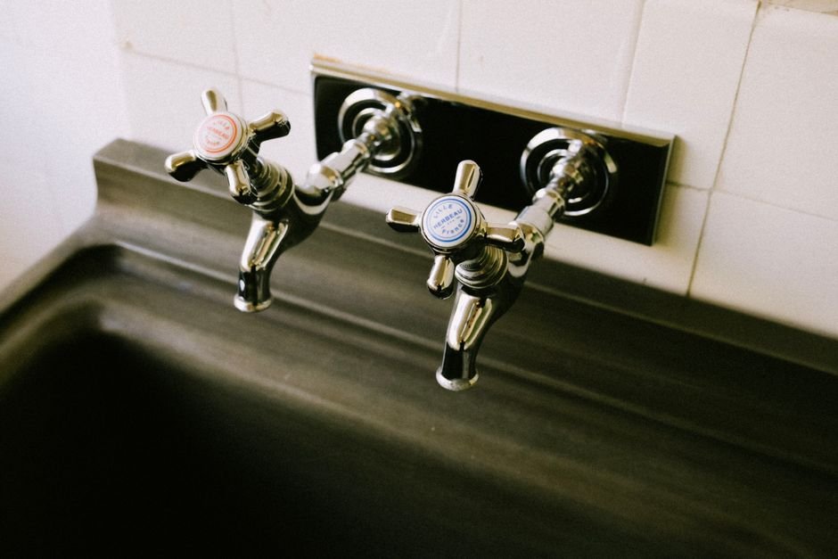 Two wall-mounted chrome cross-handle taps labelled hot and cold above a deep stainless steel trough basin, the classic utility sink faucet arrangement
