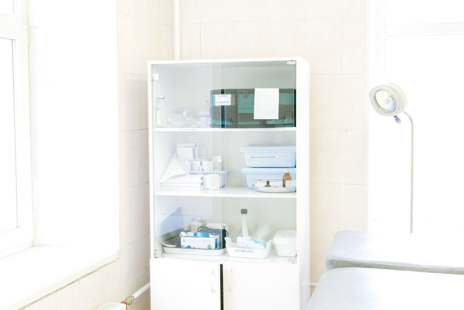 Tall white glass-fronted clinical cabinet holding trays of sterile supplies and instrument boxes in a bright tiled treatment room beside an examination lamp