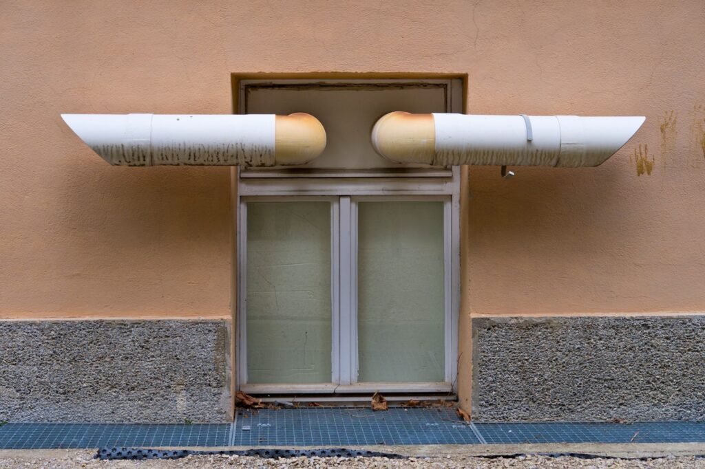 Two white exhaust ducts poking through a render wall above a basement window, lint staining streaked down the wall beneath each outlet