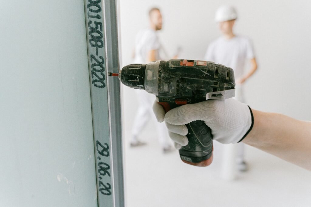 A gloved hand drives a screw with a cordless drill into the metal edge track of a plasterboard wall, with two workers out of focus behind