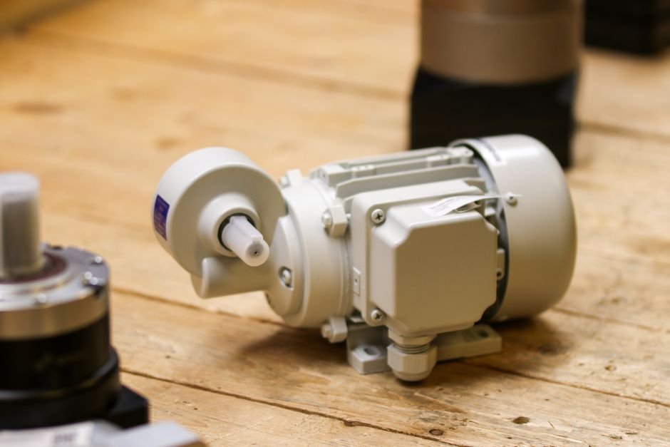 A pale grey electric motor with a bolted terminal box, mounting feet and a short output shaft, resting on a rough wooden workbench beside other motor housings in a workshop