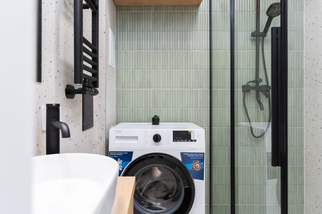 A single white front-load washing machine fitted into a narrow tiled bathroom beside a glass shower enclosure and basin